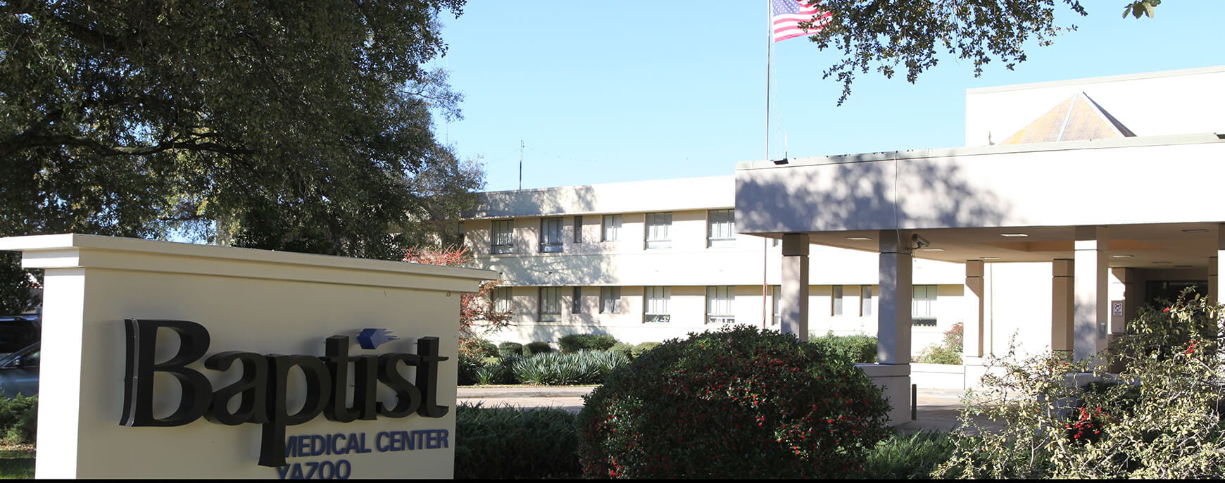 Exterior view of a Baptist Medical Center, showing the entrance canopy, landscaped grounds, and the hospital building with an American flag flying above.