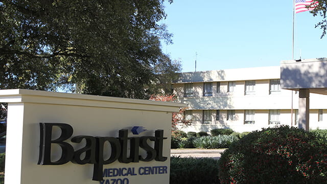 Exterior view of a Baptist Medical Center, showing the entrance canopy, landscaped grounds, and the hospital building with an American flag flying above.