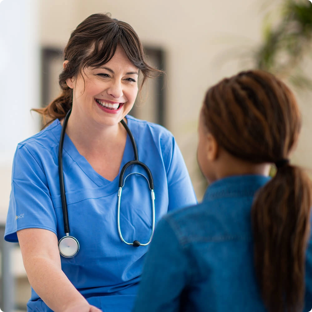 A smiling nurse in blue scrubs talks with a young girl in a denim shirt, both seated and facing each other.