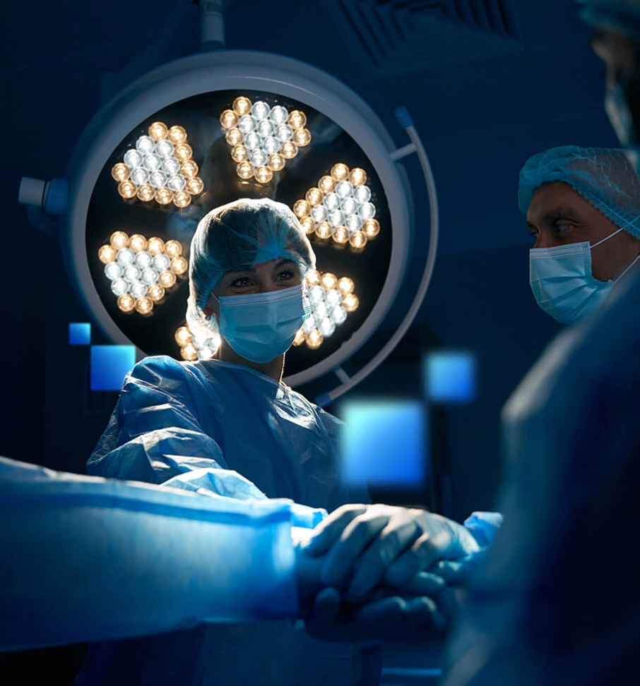 Three surgeons in masks and gowns stand under a large surgical light in a dimly lit operating room.